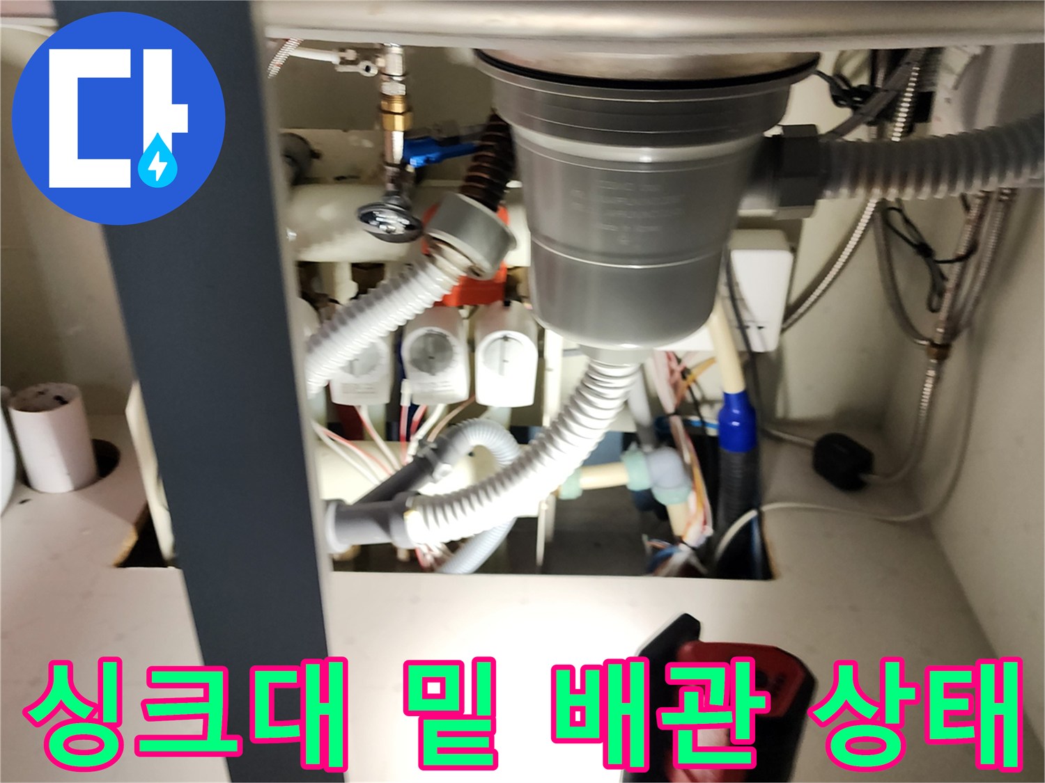 어두운 싱크대 하부장 내부의 배수 부속과 급수 라인을 촬영한 사진