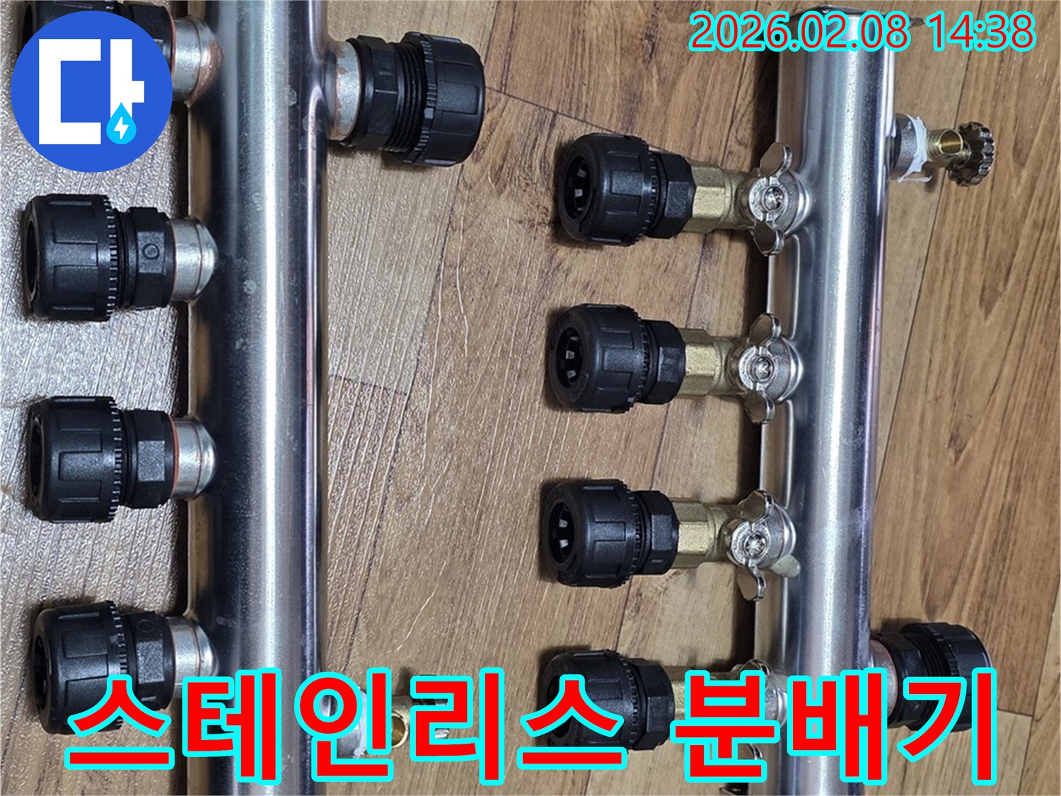 두-개의-반짝이는-스테인리스-온수분배기가-나무-바닥에-놓여-있으며-각각-여러-개의-검은색-연결-단자와-금색-밸브를-포함하고-있습니다