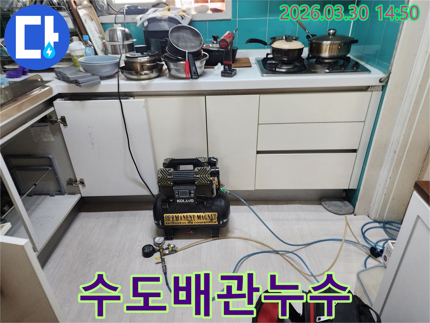 주방 하부장 아래에 위치한 수도 배관에-누수 탐지용 공압 장비가 연결되어-압력 테스트가 진행되고 있습니다.