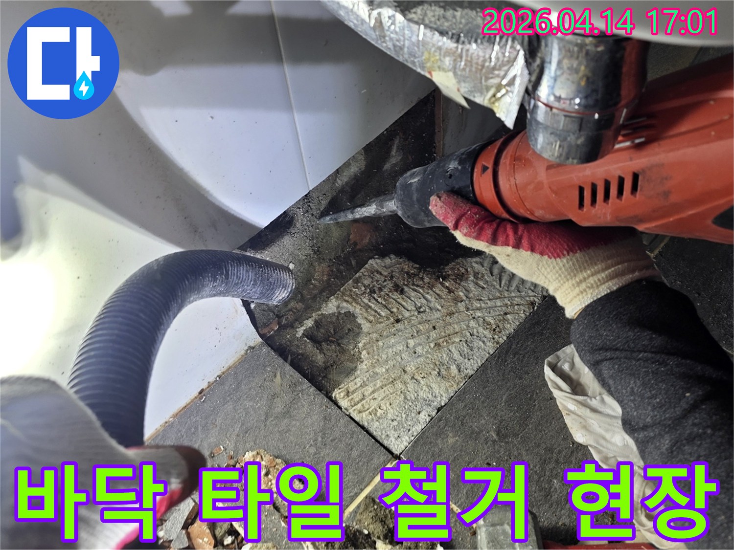누수 탐지 후 바닥 타일을 철거하는 작업자가 드릴을 사용하며-파손된 타일 조각과 흡입 호스가 보이는 공사 현장