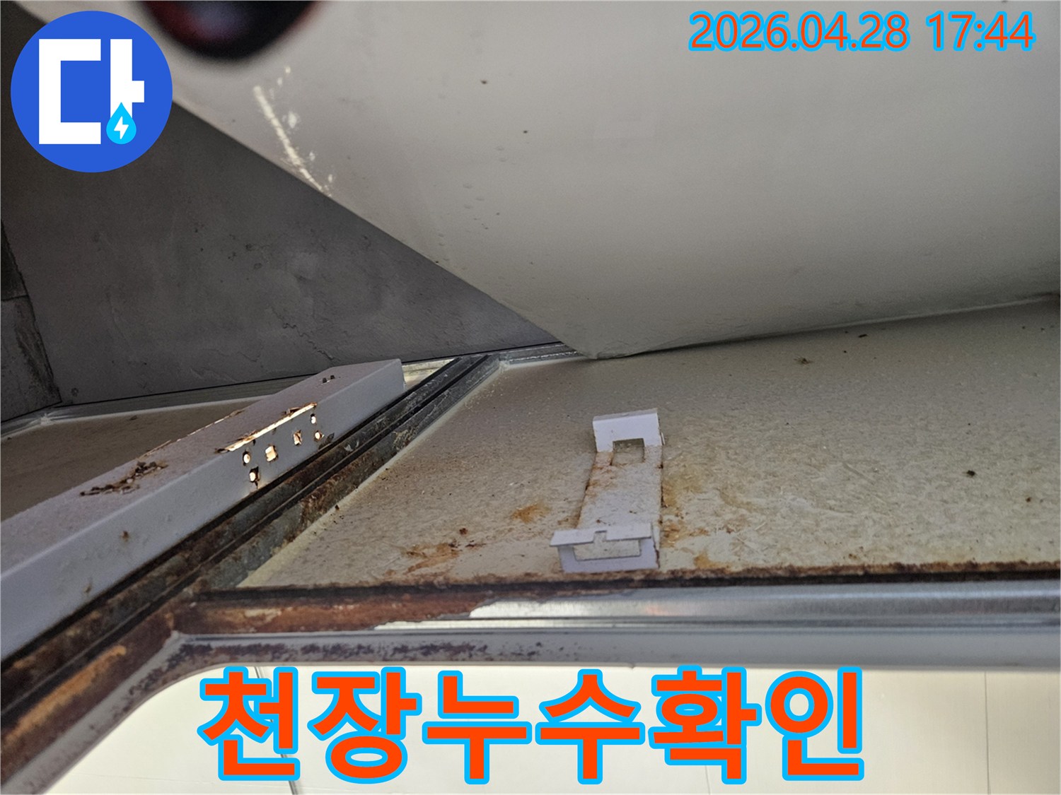 아래층 욕실 천장의 누수 흔적을 보여주는 사진입니다-녹슨 금속 구조물과 변색된 천장 마감재가 명확하게 보입니다.