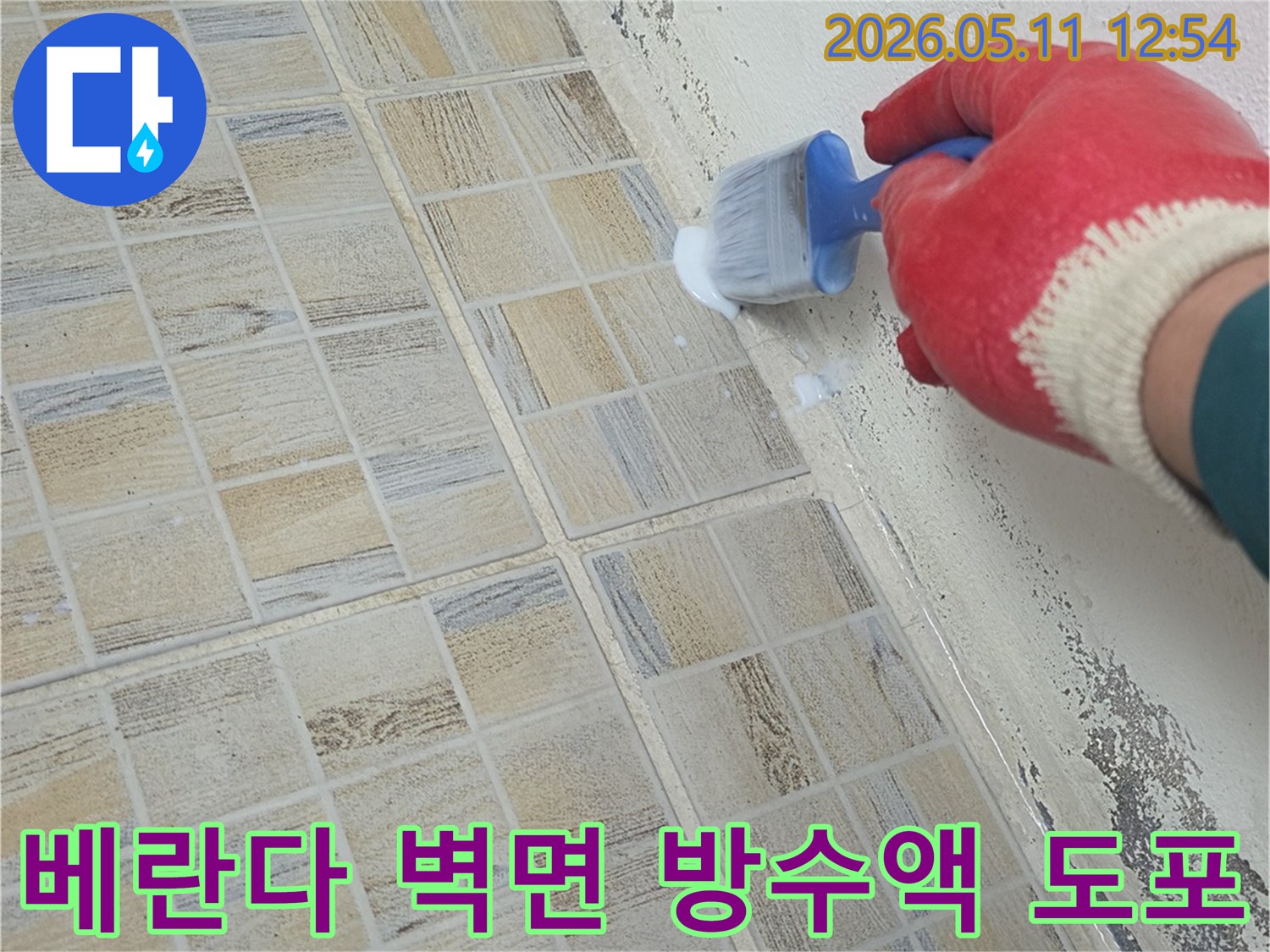 붉은 장갑을 낀 작업자가-베란다 벽면 하단과 타일 경계에-방수액을 붓으로 꼼꼼히 바르며-습기 문제를 해결하고 있습니다