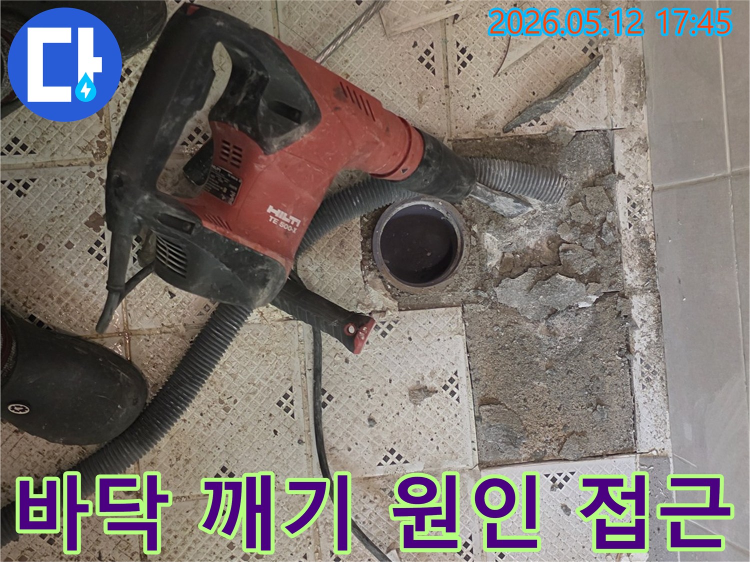 누수 원인에 접근하기 위해-바닥 타일을 깨는-강력한 드릴 장비와-작업자의 발이 보이는-현장 사진입니다.