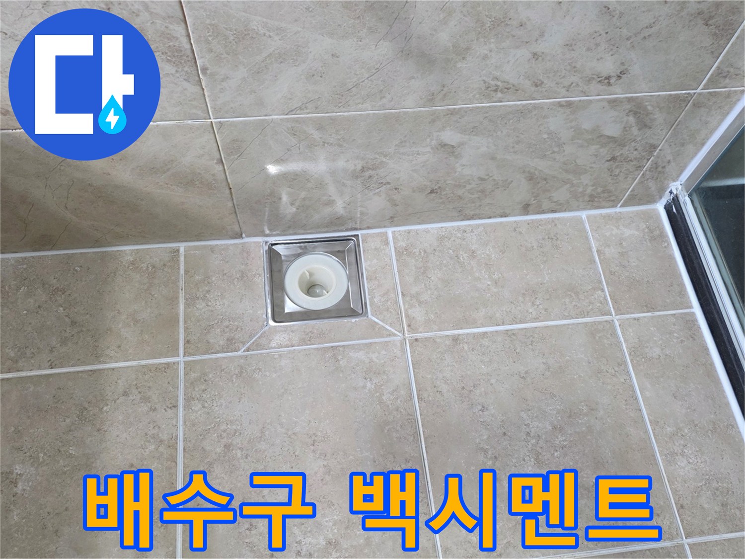 베이지색 타일 바닥에 설치된-사각형 금속 배수구의 모습으로-주변 백시멘트의 마감 상태를 보여줍니다