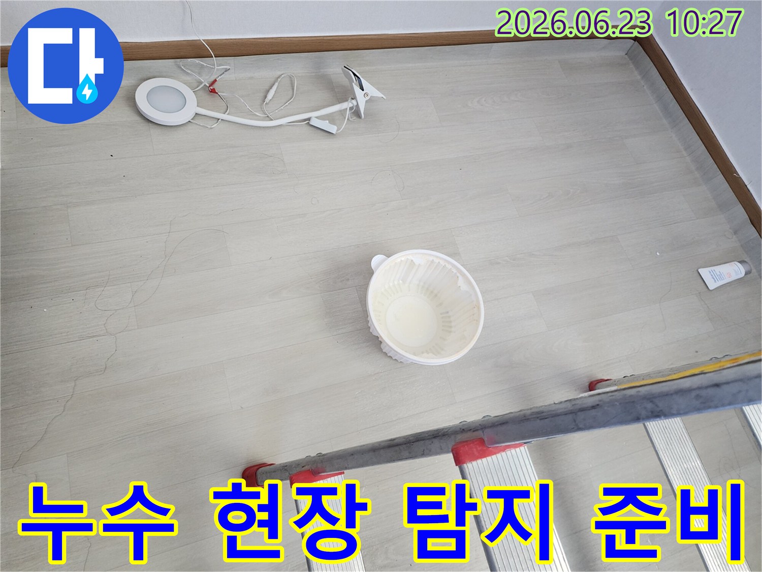 누수로 인해 바닥에 떨어진 물을 임시로 받는 그릇과-누수 지점 확인 및 수리를 위한 사다리-작업용 조명 장비가 바닥에 놓여 있는-누수 공사 전 현장 모습입니다