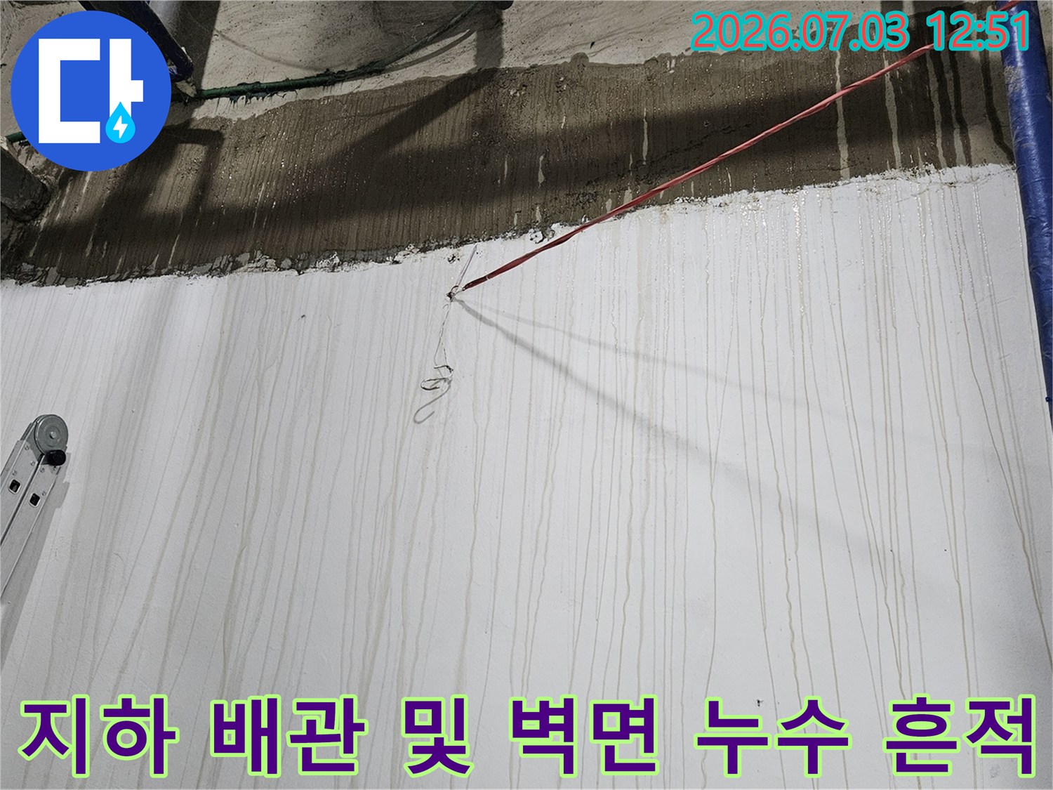 파란색 수직 배관과 그 옆으로 물이 흘러내린 자국이 가득한 흰색 지하 벽면-왼쪽에 사다리 일부가 보임