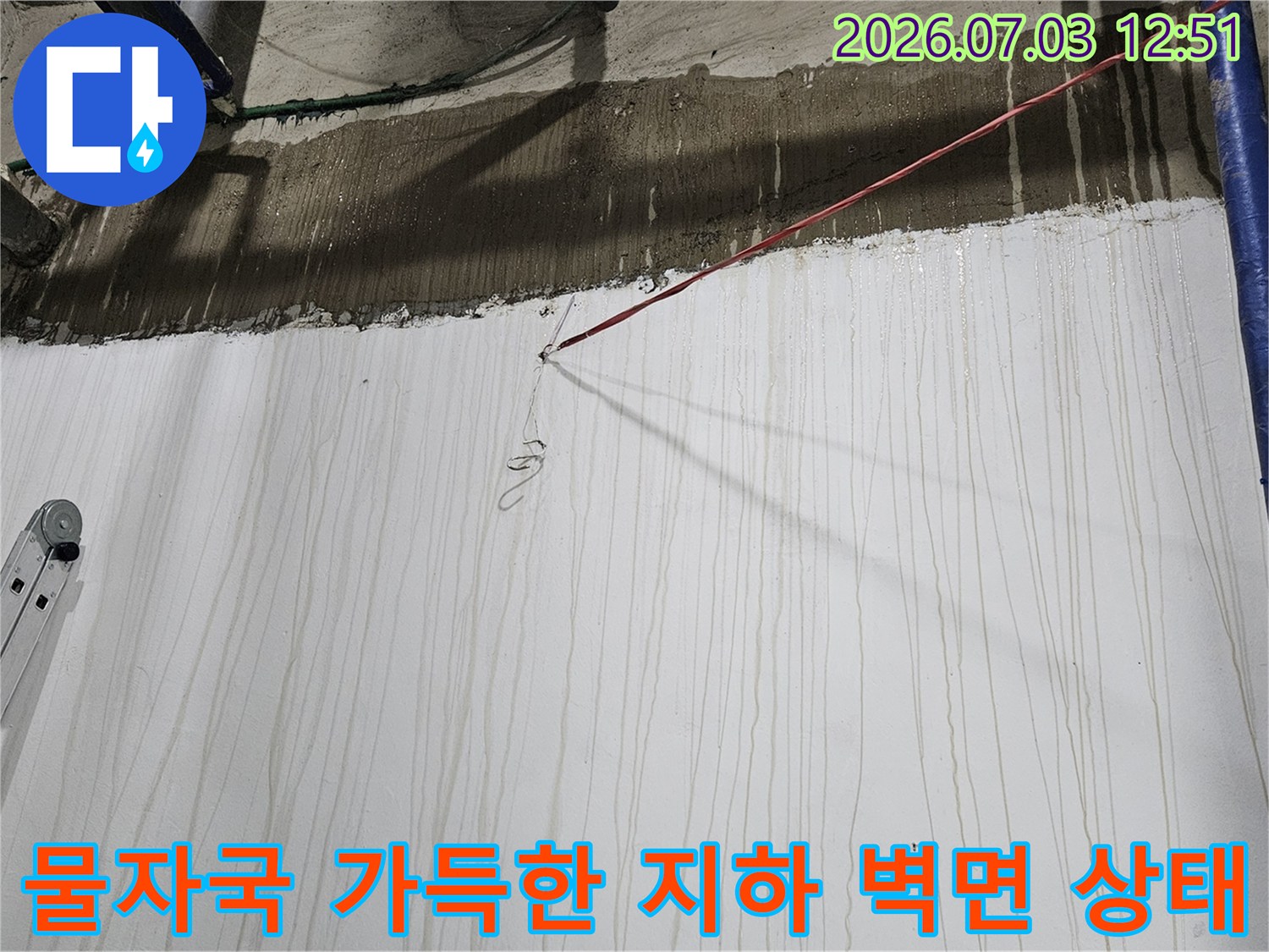 흰색 페인트 벽면에 세로로 길게 형성된 누수 오염 자국-우측에는 파란 배관이 수직으로 설치됨
