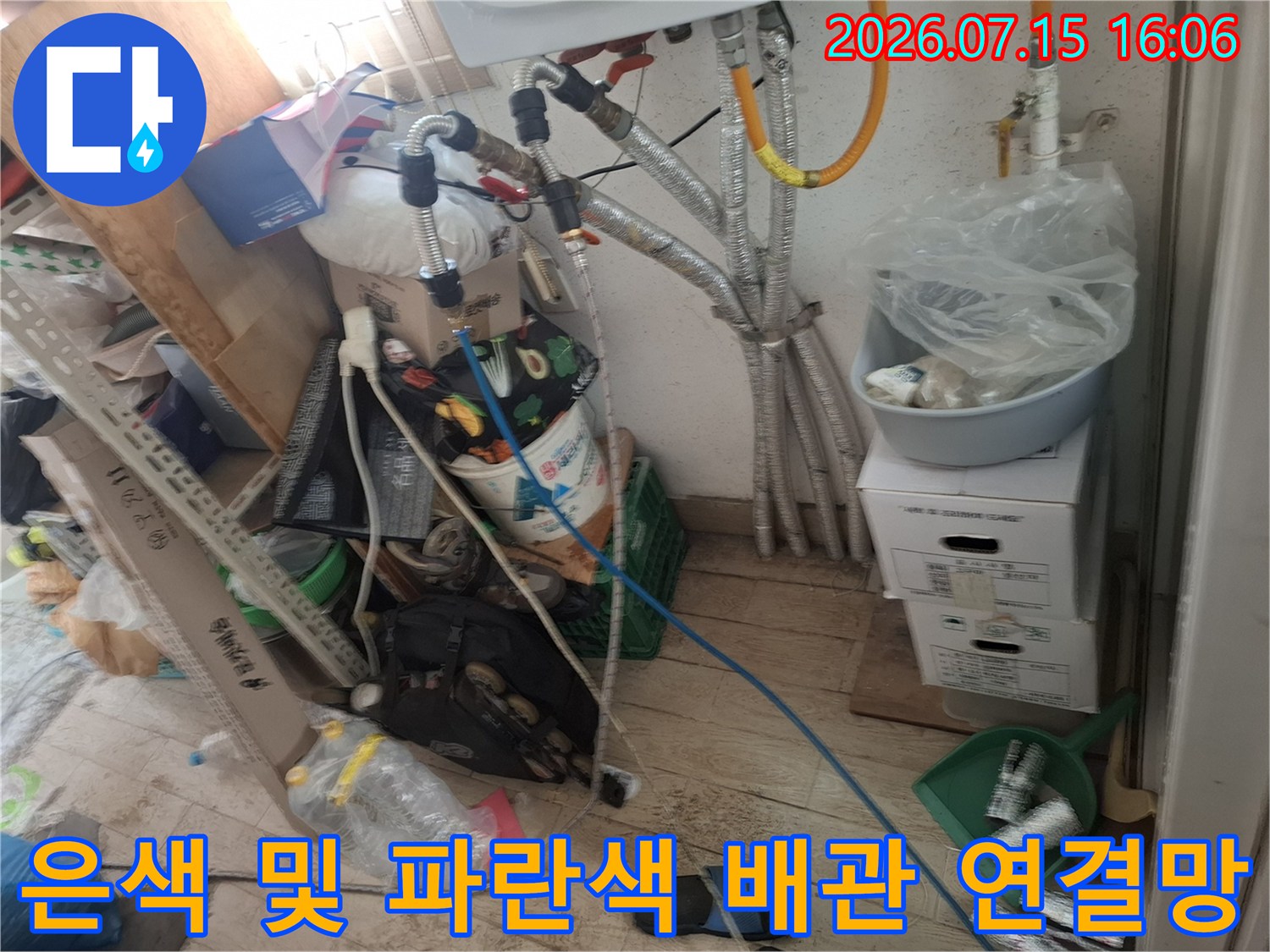 보일러 본체 아래로 설치된 은색 금속 주름관-파란색 플라스틱 호스가 교차 연결된 모습