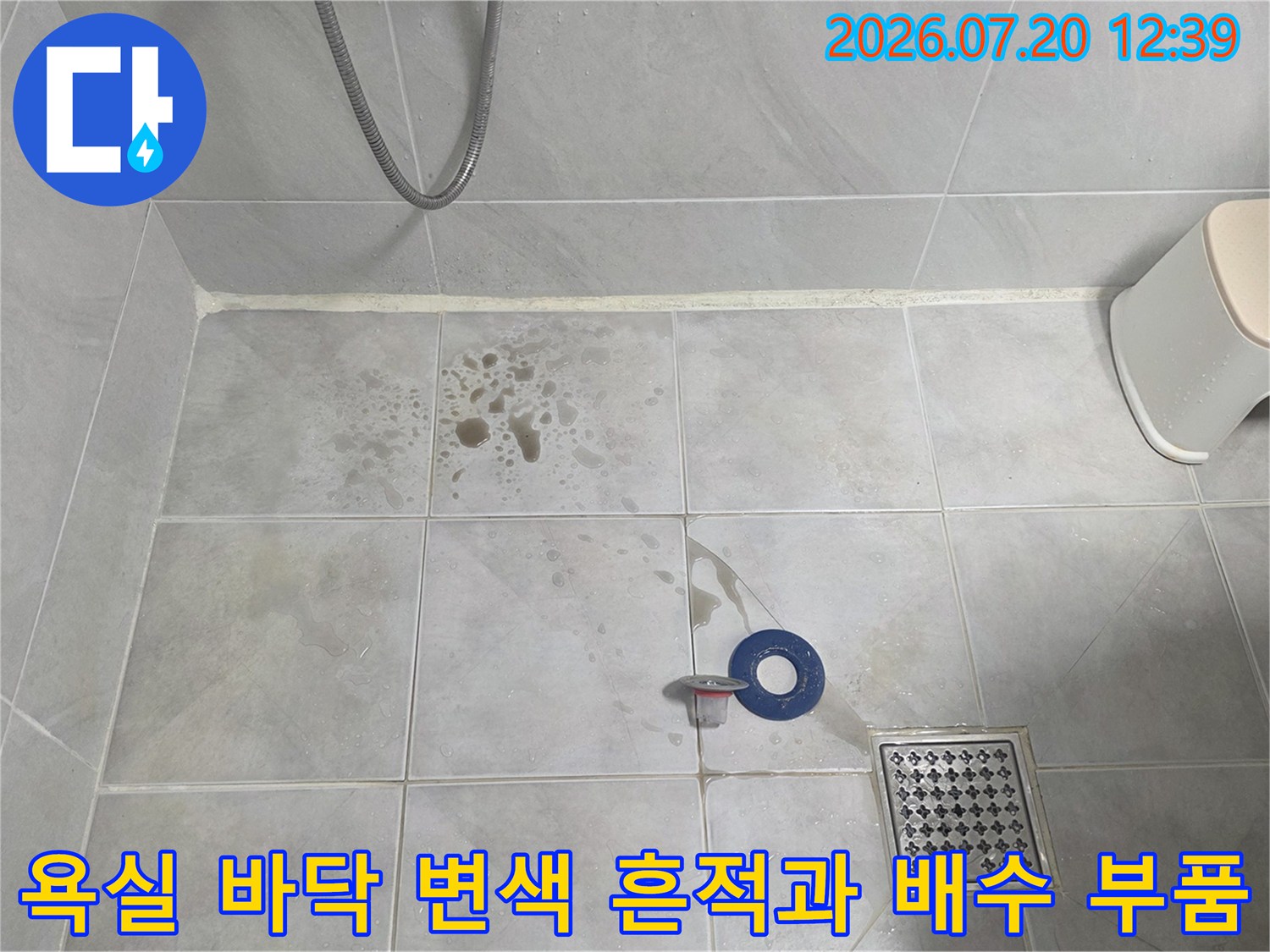 흰색 욕실 타일 위에 갈색의 습기 얼룩이 있고 배수구 주변에 파란색 링 부속이 있는 사진