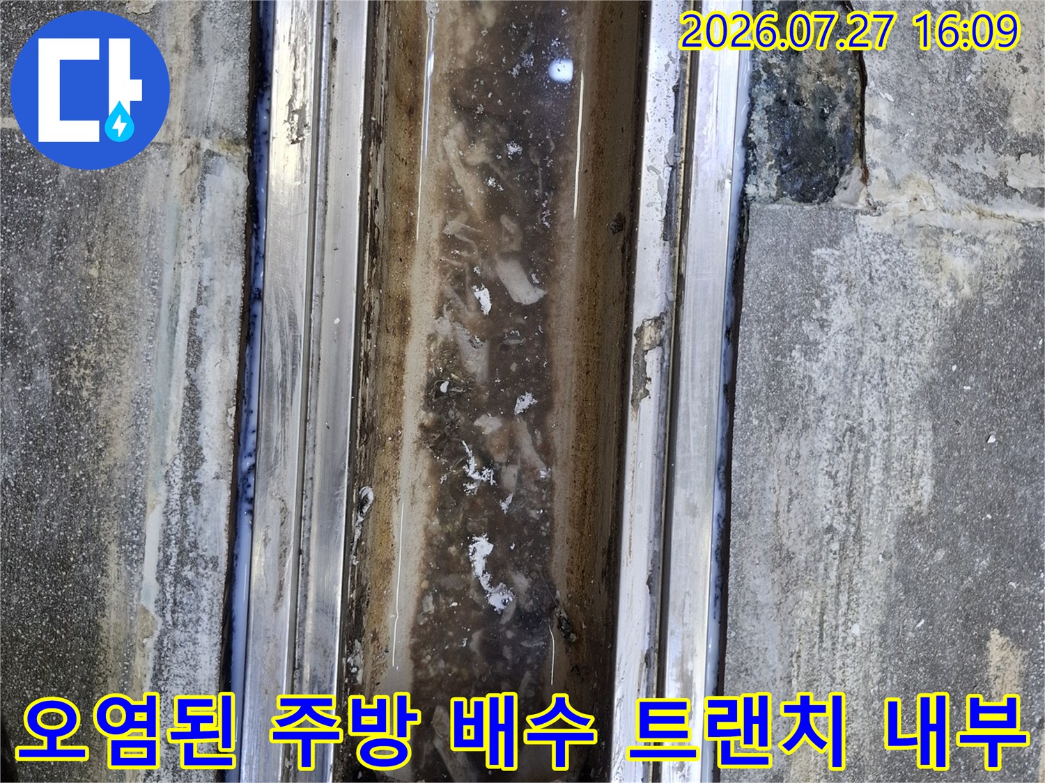 주방 바닥에 설치된 금속제 배수 트랜치의 덮개가 제거되어 내부의 오염물과 균열이 드러난 모습