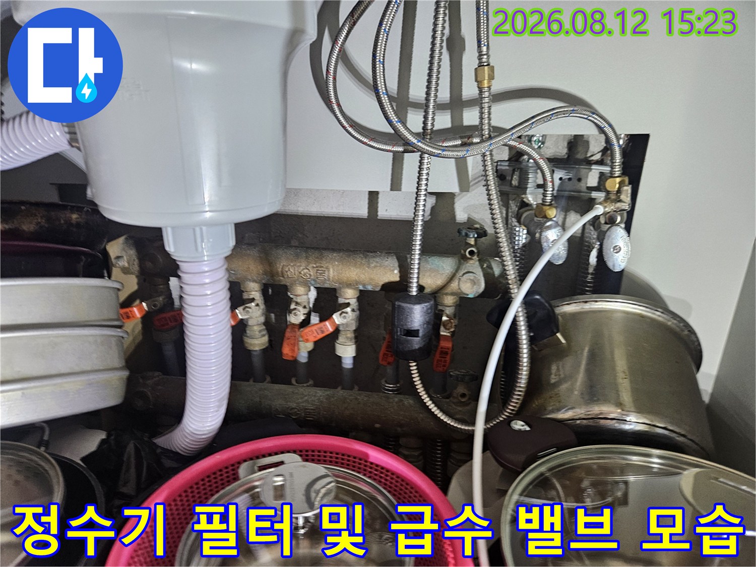 주방 하부장 내부의 흰색 정수 장치와 금속제 수도 배관들이 연결된 복잡한 설비 구조