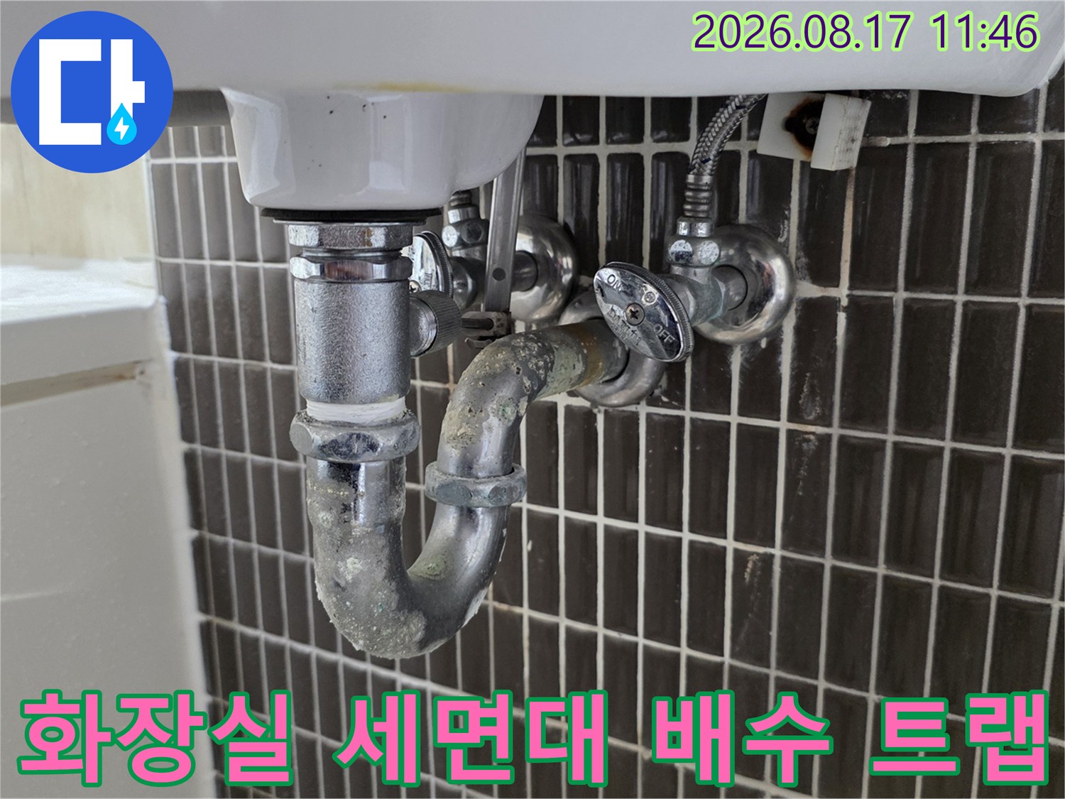 세면대 하부의 금속 재질 배수 트랩과 벽면 타일 부위 모습입니다.