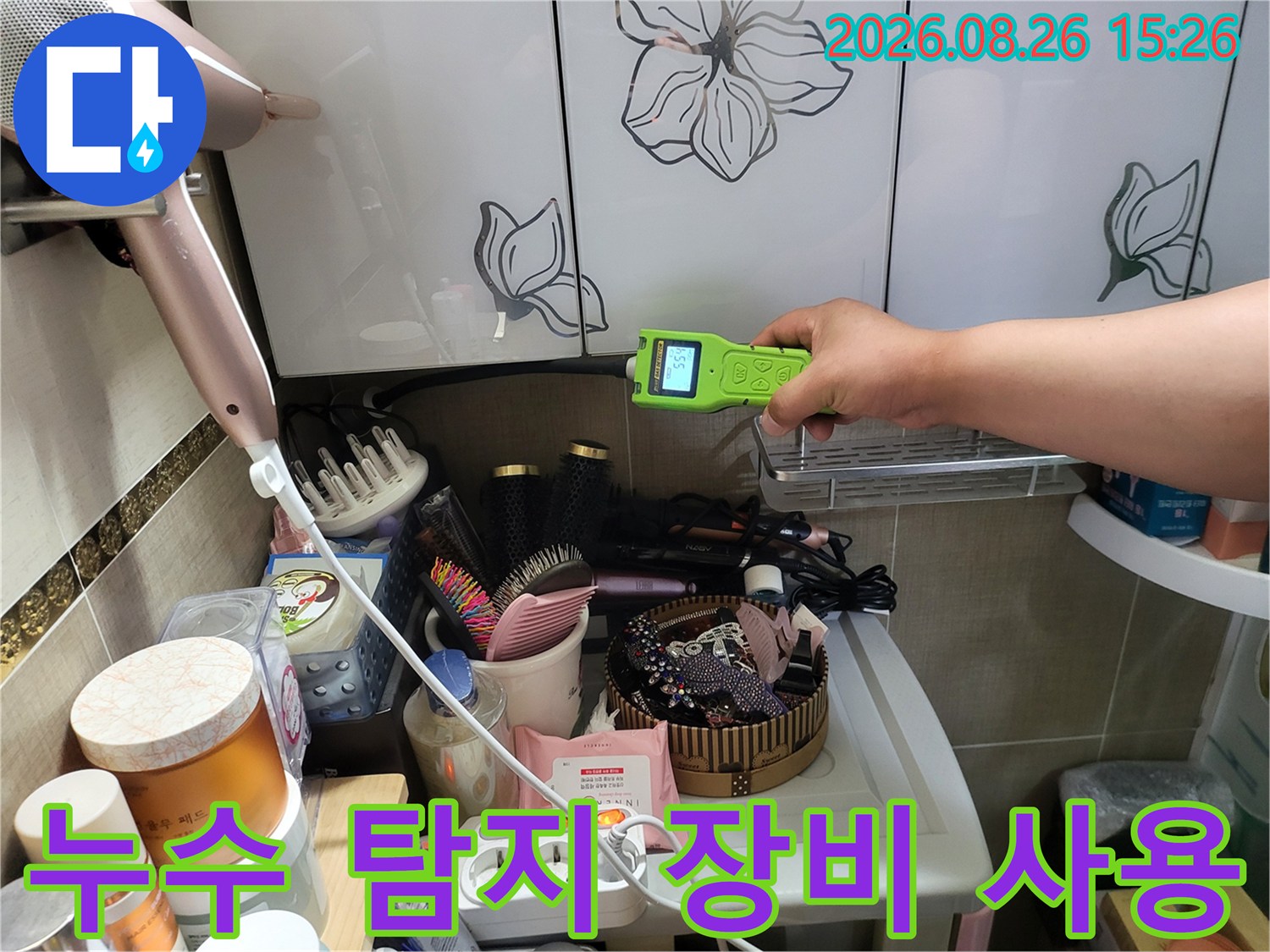 화장실 선반 아래 공간에서 누수 여부를 확인하기 위해 휴대용 측정기를 조작하고 있습니다.